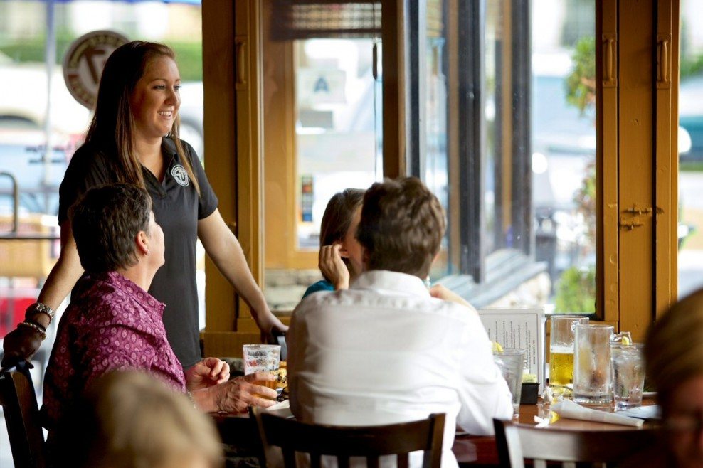 Server chatting with guests at a window table inside Burger Tavern 77